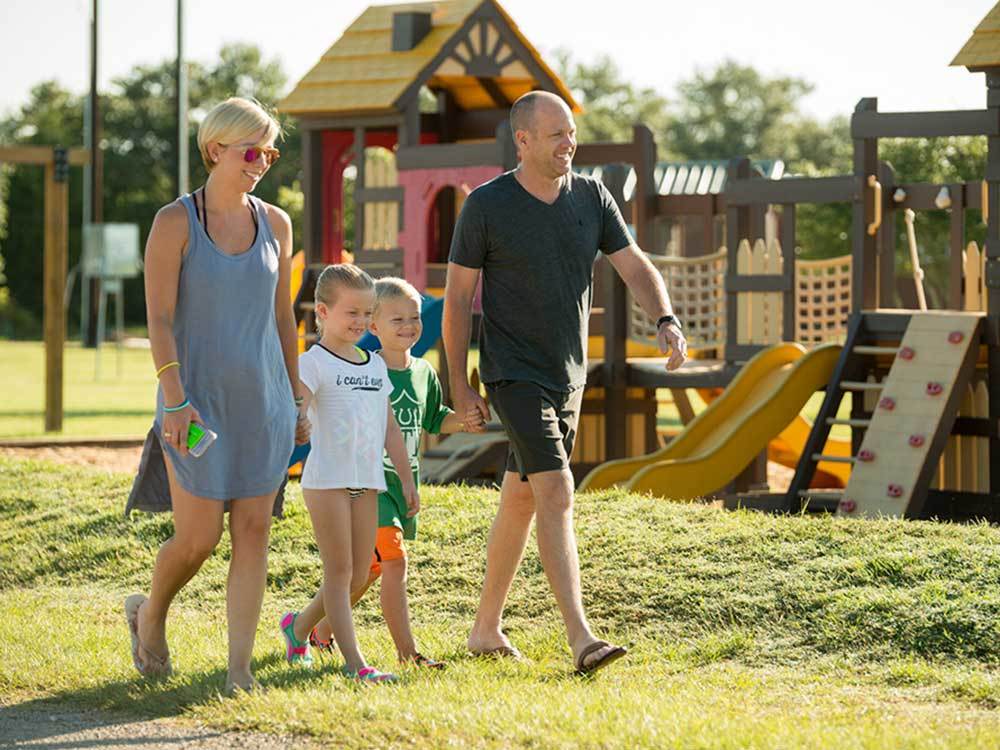 Two kids flanked by their parents walk hand-hand-in-hand past some play structures.