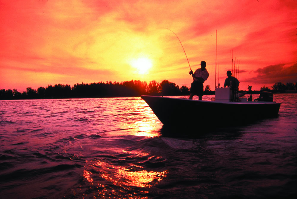 An angler stands in a boat as he pulls on a fish, his rod bent by the strain.