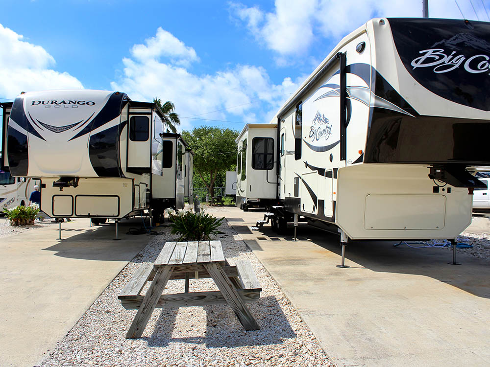 A picnic bench sits between two concrete camping spaces occupied by two large fifth-wheels.