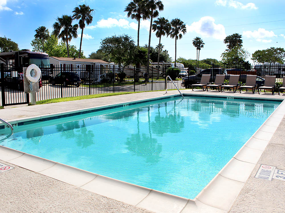 A pool with aqua-marine water, deck chairs overlooking one side and a black iron fence with pam trees and RVs in background.