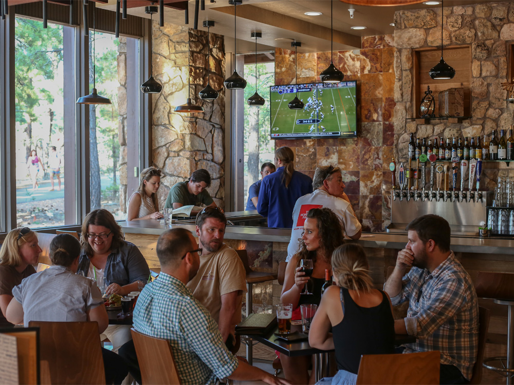 Patrons of a restaurant chat around a pair of tables with a bar in the background as a TV mounted on the wall shows football.