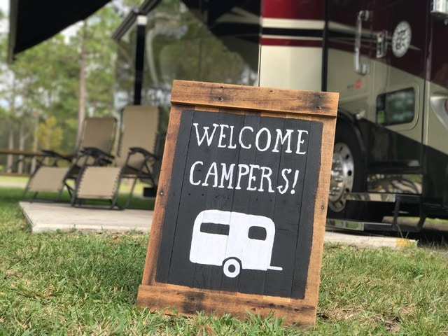 A "welcome campers" sign -- white letters against black background -- propped up with motorhome in background.