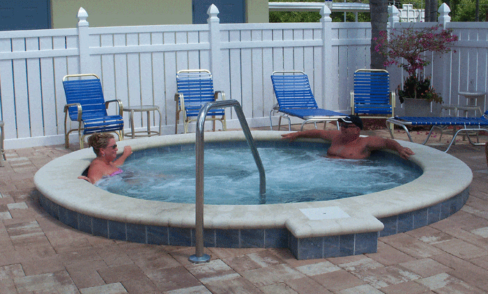 A man and woman sit in a hot tub with chaise lounges on the surrounding brickwork and white fence in background.