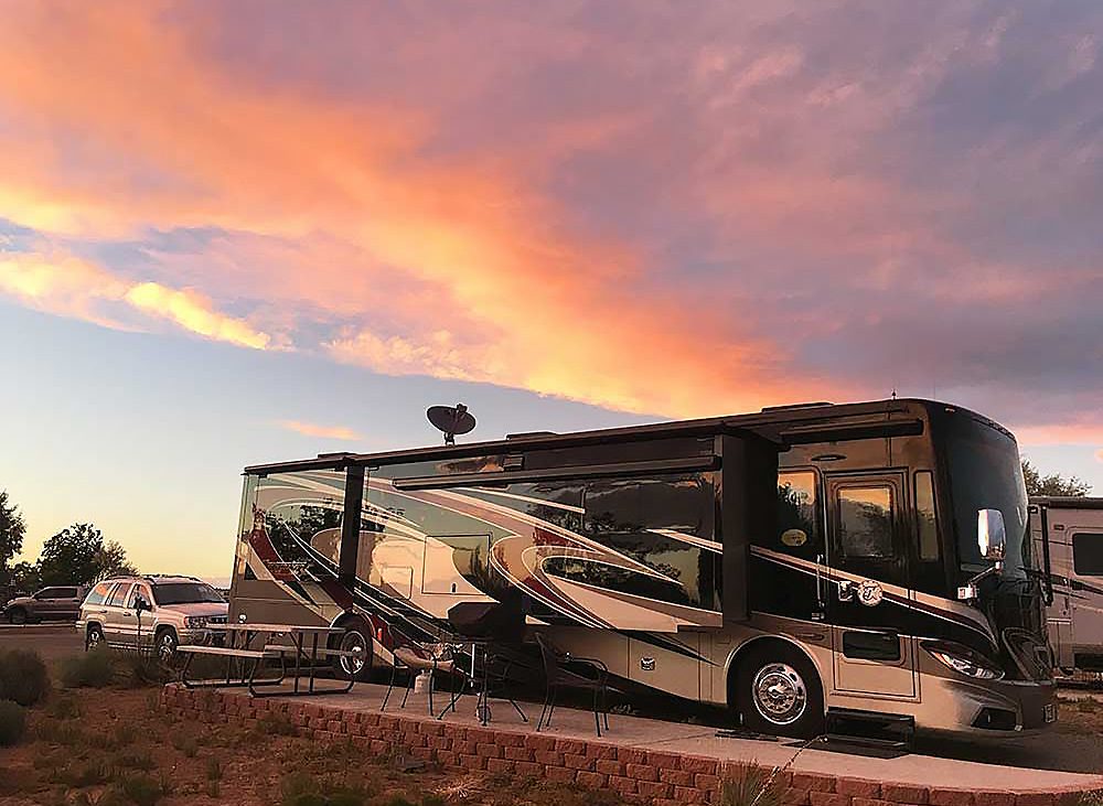 A motorhome with brown and tan graphics against a sunset sky with light burning red on low hanging clouds.