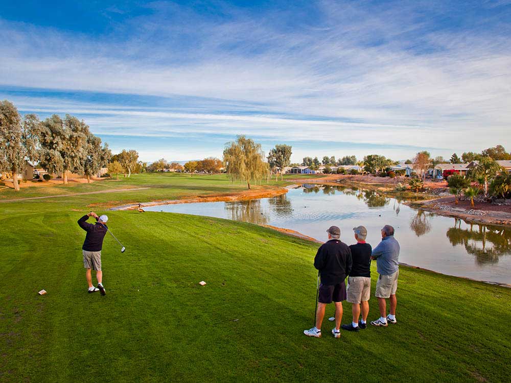 A man hits a golf ball down a lush fairway from the tee with a pond to the right — three men look on.