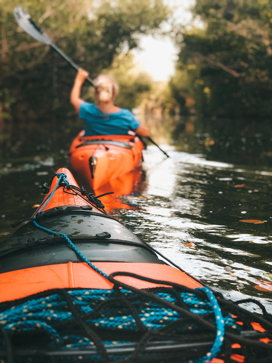A woman paddles a kayak through a narrow channel bordered on each side by dense foliage.