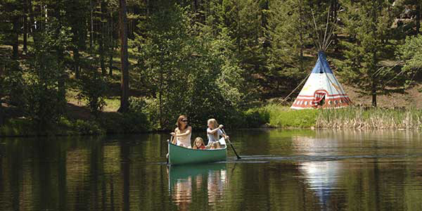 Two women and a child paddle a green canoe with tepee on bank.