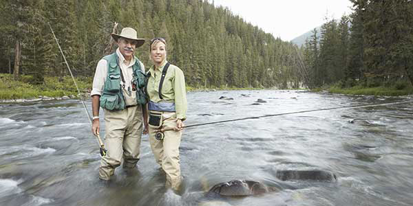 A man and woman wade in a rushing stream with fishing poles.