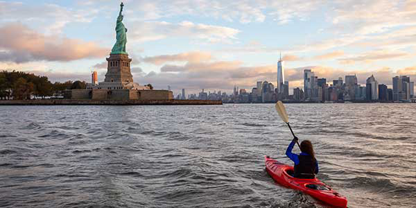 A woman paddles a red kayak toward the Statue of Liberty.