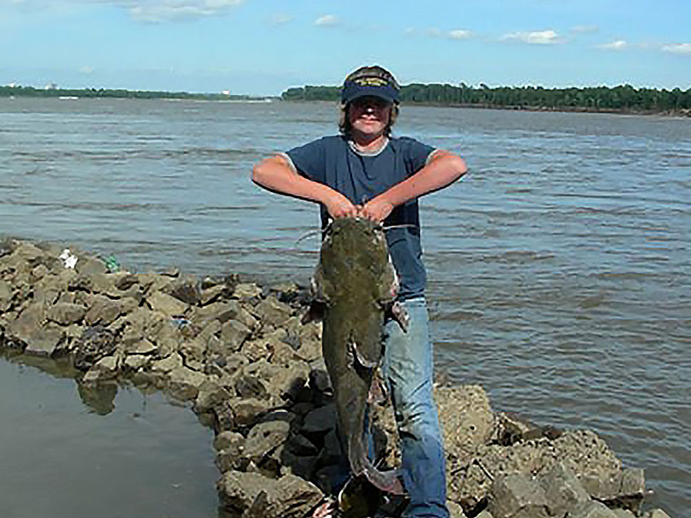 A boy holds up a huge catfish on a jetty on the river.