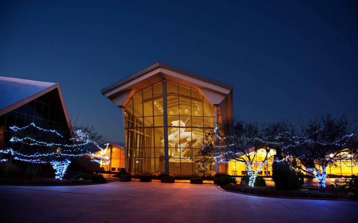 Large building with floor to ceiling windows displaying horse sculpture at night with Christmas lights