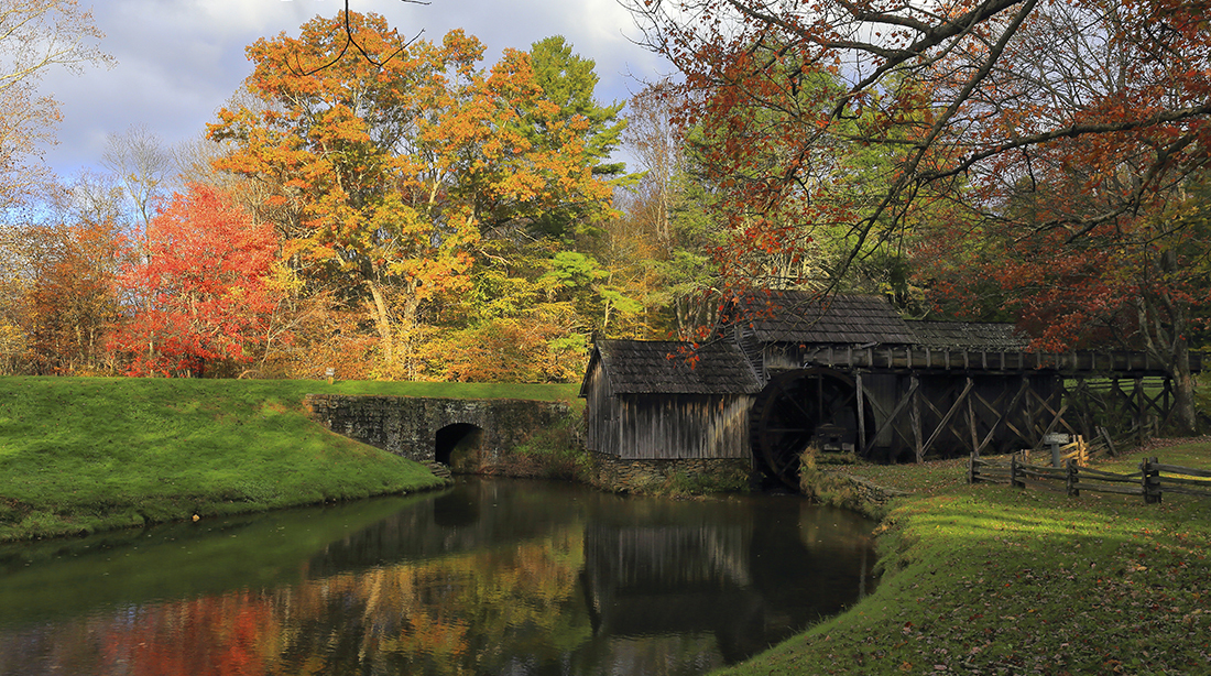 A mill on a grassy bank overlooking a stream amid fall foliage.