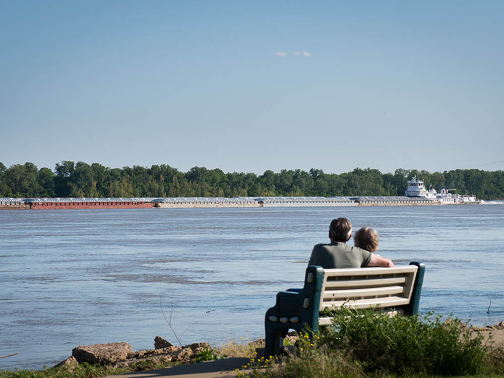 A couple on a bench watch a barge churn past.