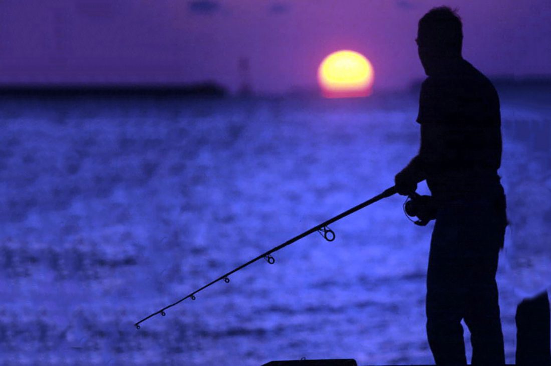 An angler stands at water's edge at sunset.