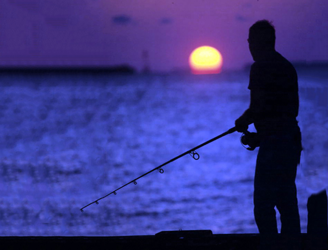 An angler stands at water's edge at sunset.