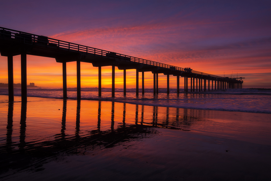 Brilliant evening sunset and silhouette of Scripps Pier with reflections on La Jolla Shores beach in California