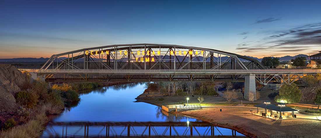 A steel bridge spans a calm river at sunset.
