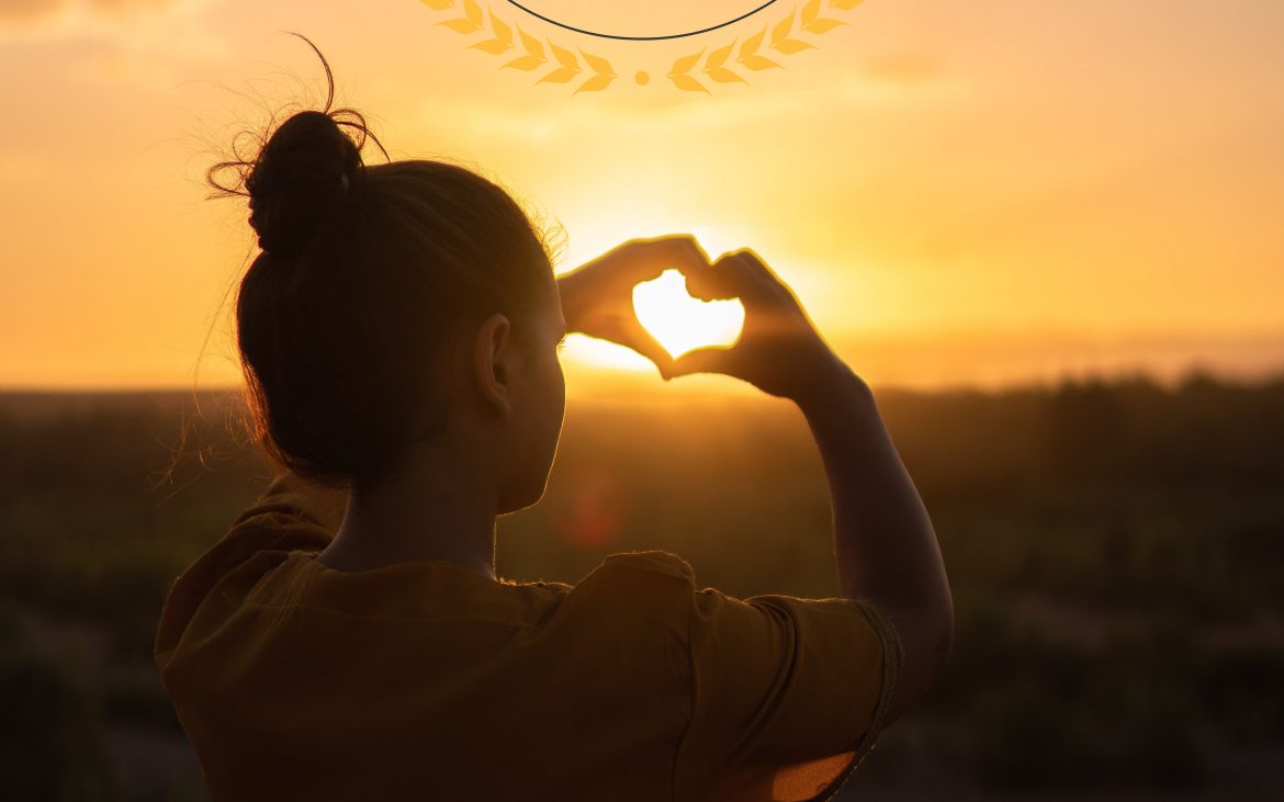 Woman makes a heart sign to a sunset sky.