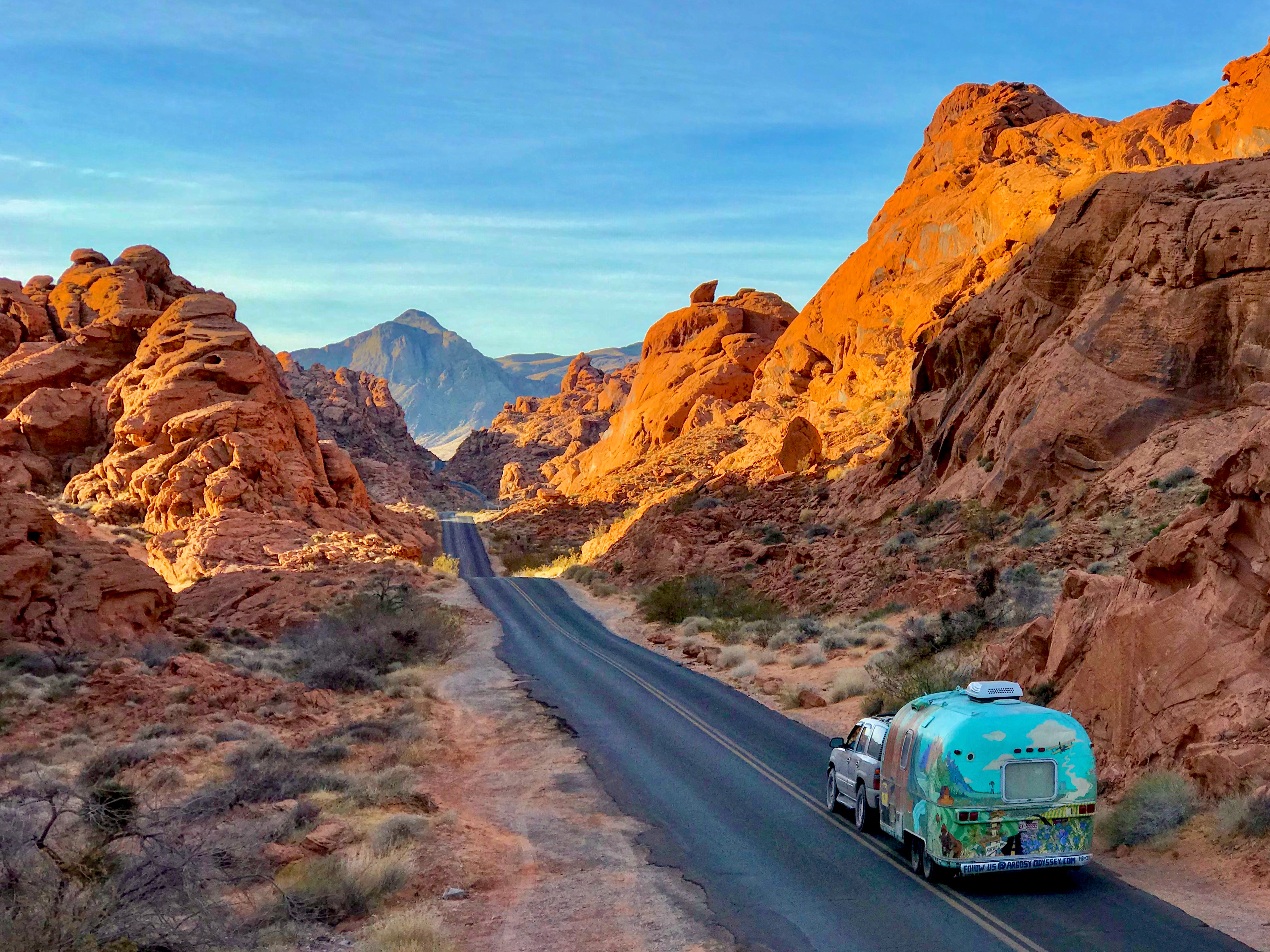A colorful Airstream trailer is towed on a highway lined with red rock formations.