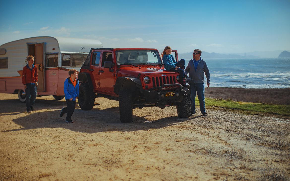 A family gathered around a red jeep hitched to a trailer.