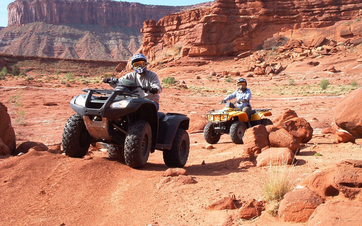A pair of ATV riders follow a dirt trail through a rugged landscape.