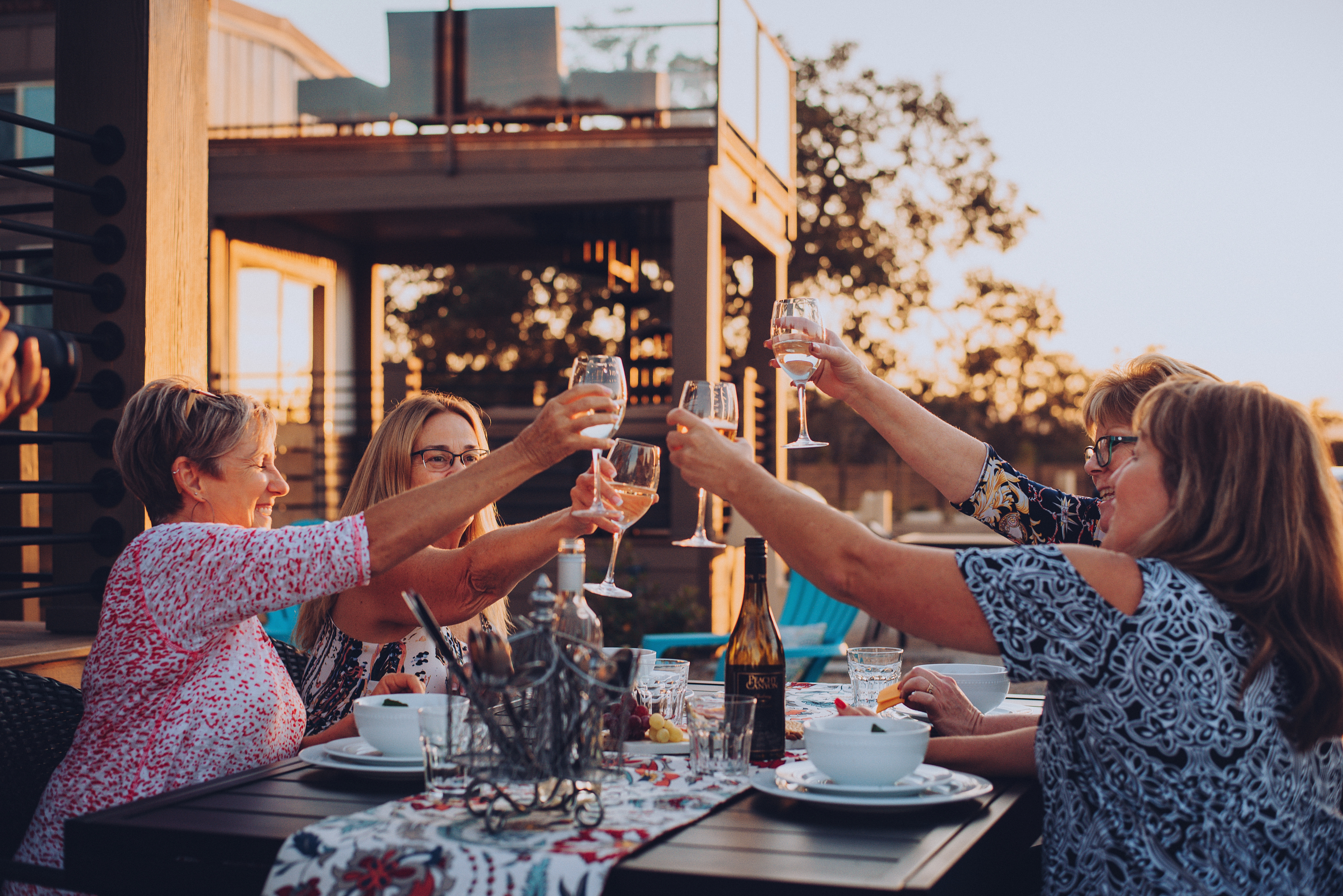 Four ladies raise glasses of white wine for a toast on an outdoor patio.