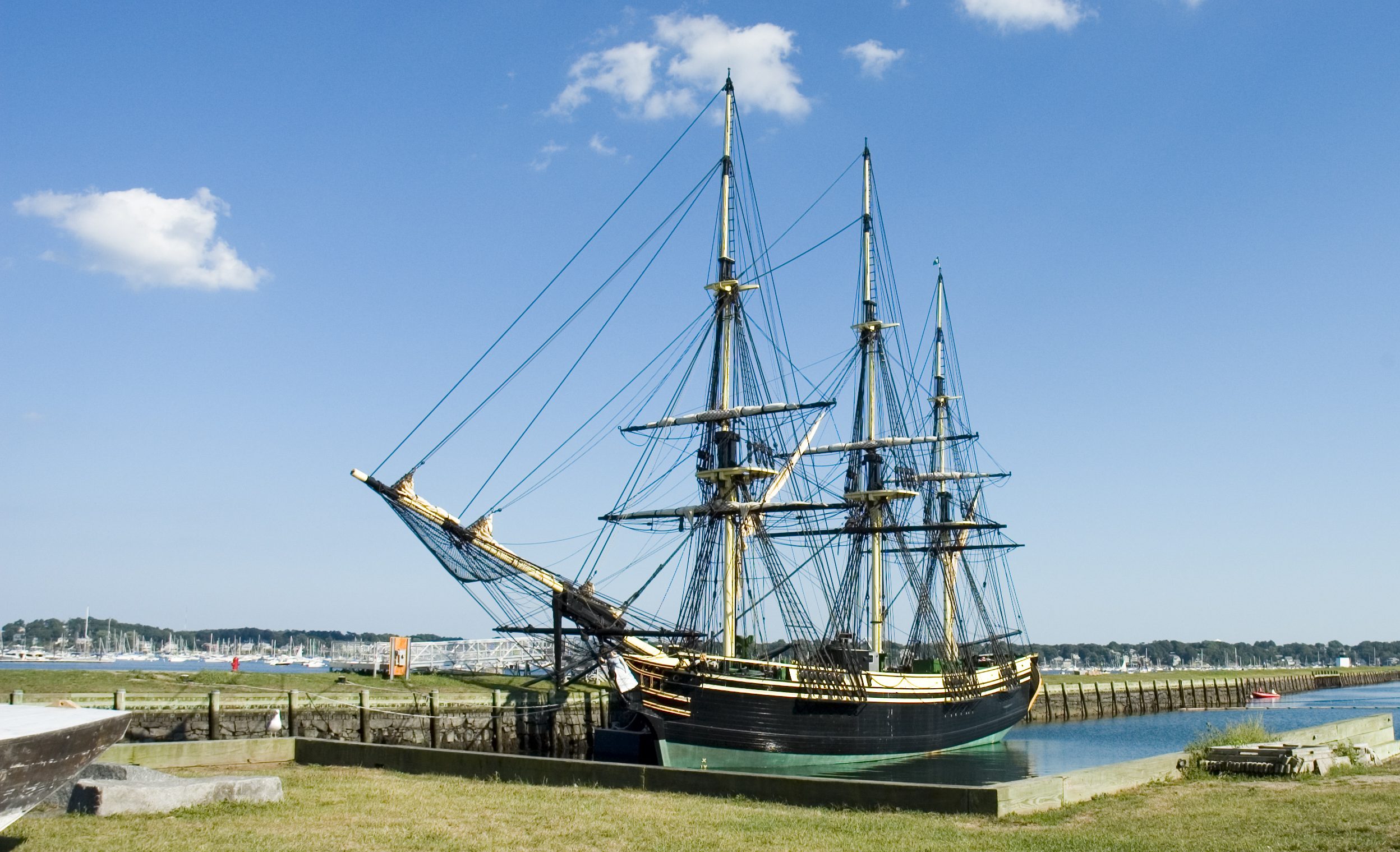 Three-masted 18th-century ship moored in a slim harbor.