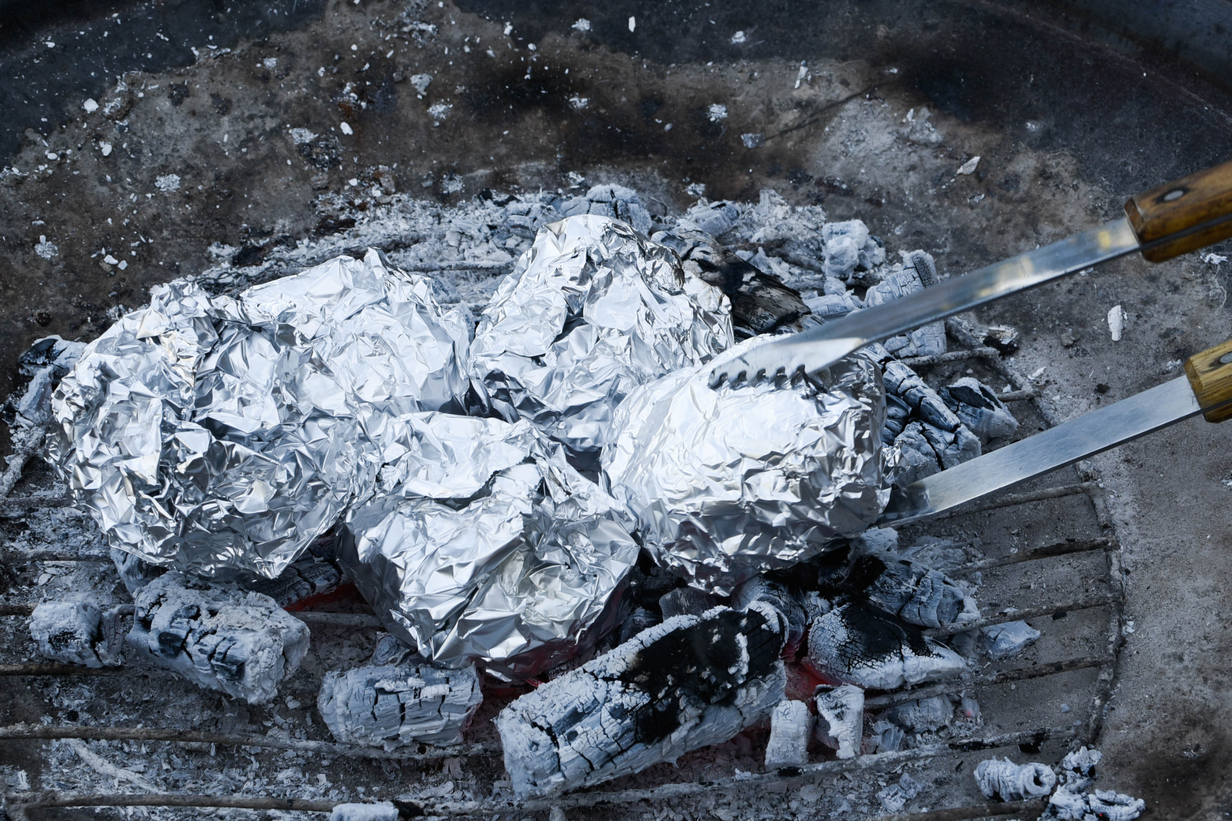 Tongs place foil-wrapped sandwiches in a barbecue pit.