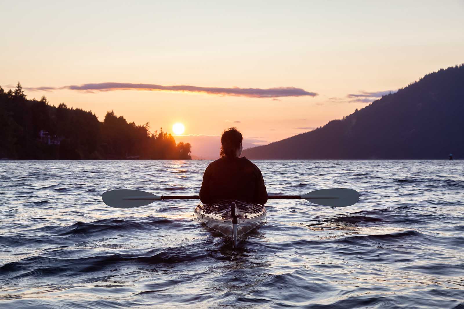 A kayaker paddles toward sunset on a lake.
