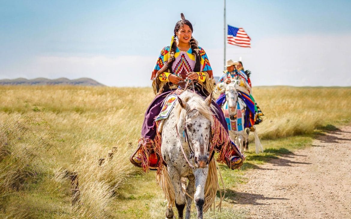 Nez Pierce of Lewis Clark Valley —A woman in tribal dress rides a white horse on a trail.