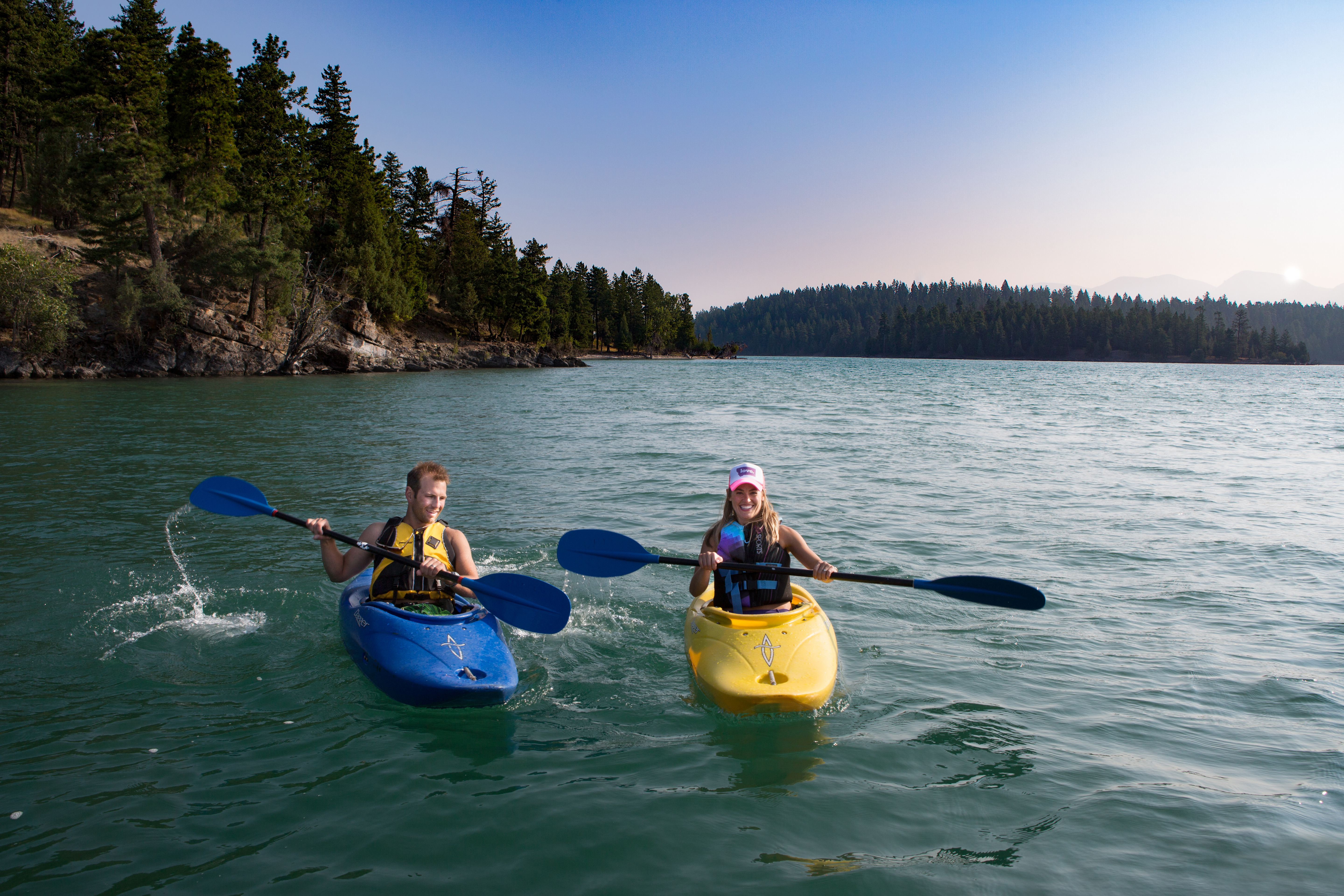 Two kayakers smile as they paddle a lake fringed in thick trees.