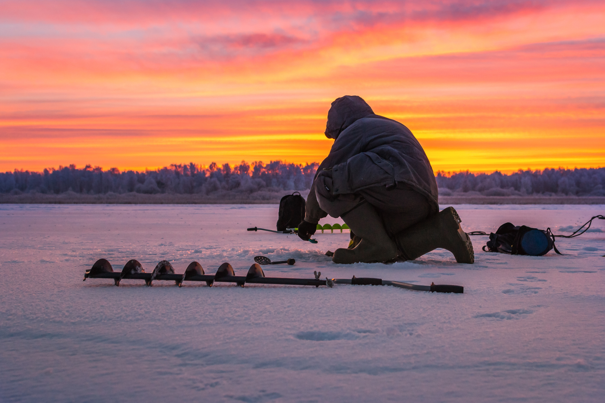 A man with augers on the ice of a frozen lake during sunrise.