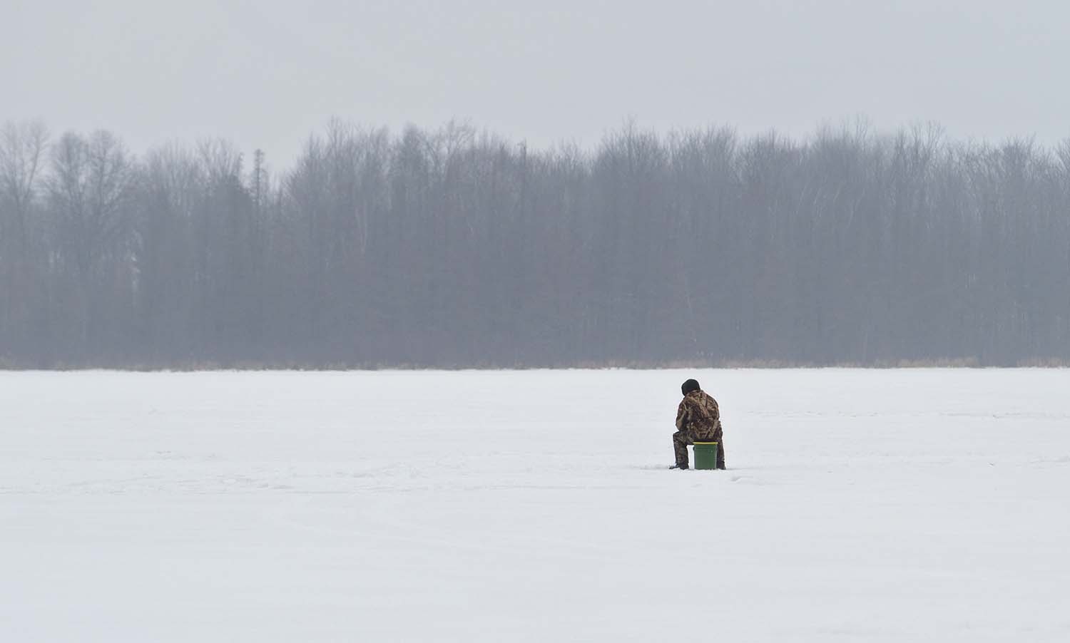 Person sitting on a bucket, ice fishing on a frozen lake in the middle of winter. Wisconsin pastime