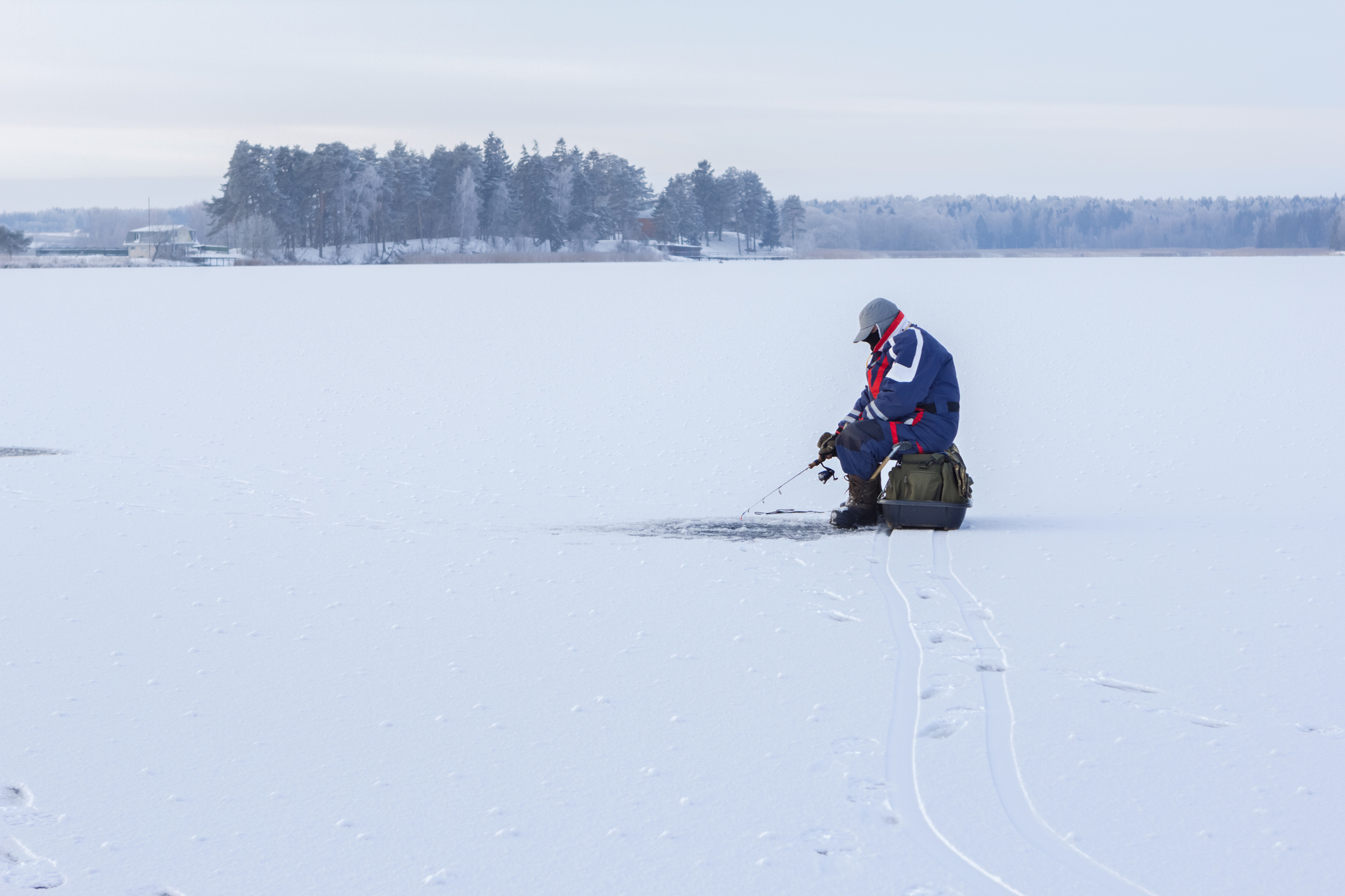A lone angler sits on the ice with a line dangling through frozen lake surface.