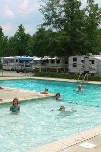 Campers swimming in a pool with RVs in background.
