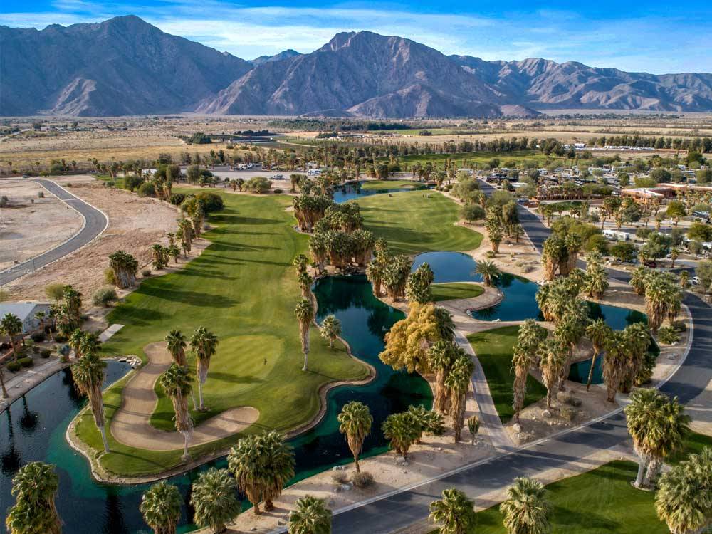 An aerial view of a golf course and RV resort against stark desert mountains.