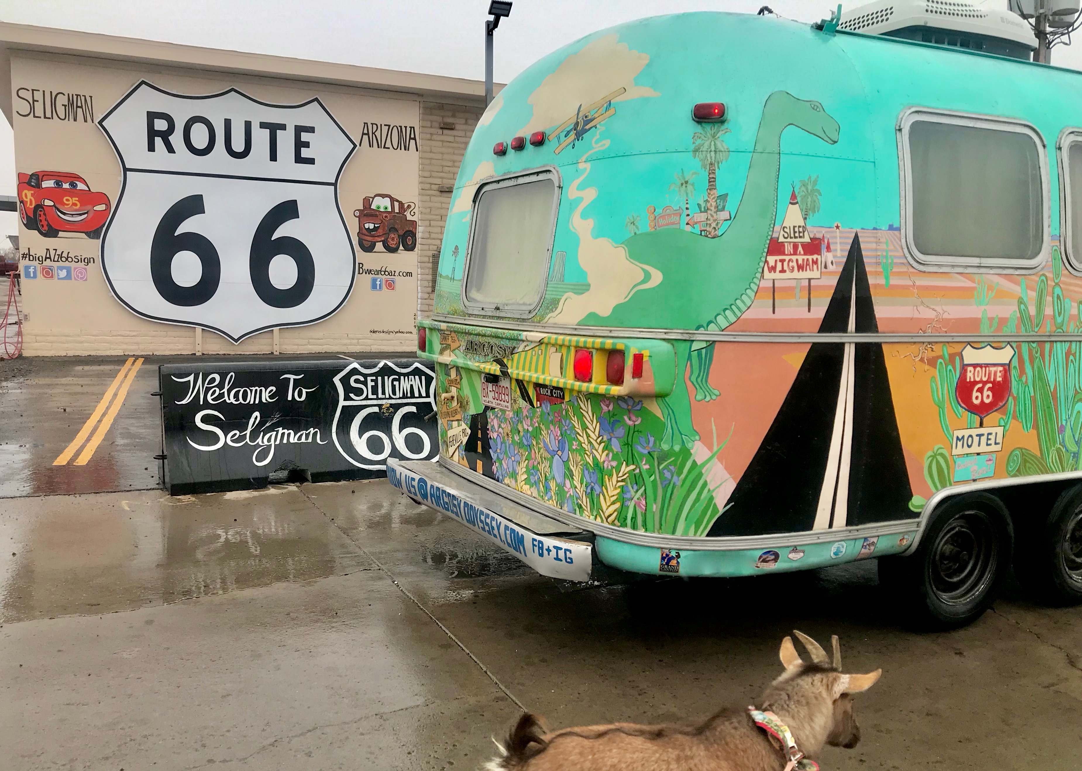 A colorful Airstream trailer parked near a Route 66 sign with goat in foreground.