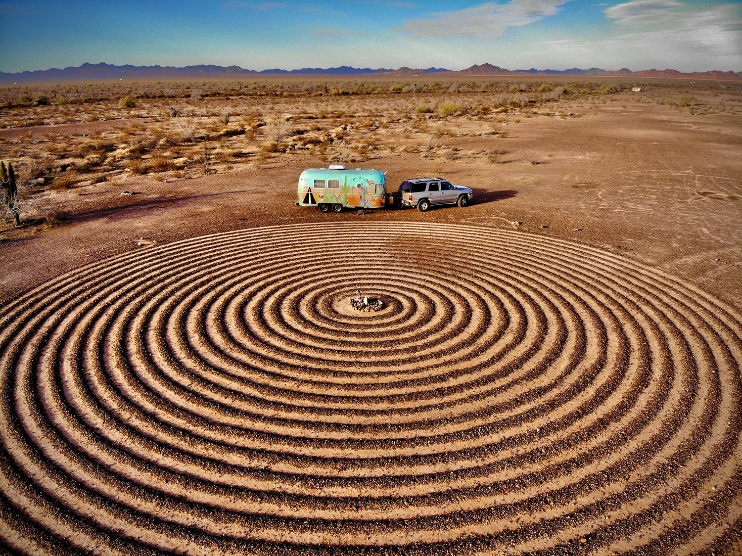 SUV with airstream trailer parked near a spiral labyrinth carved into the earth in a desert.