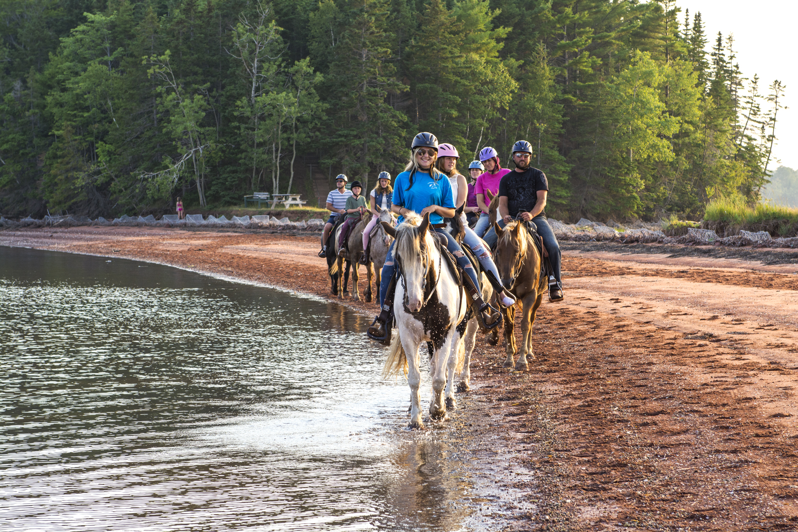A group of young people riding horses along a lakeshore with tall trees in background.