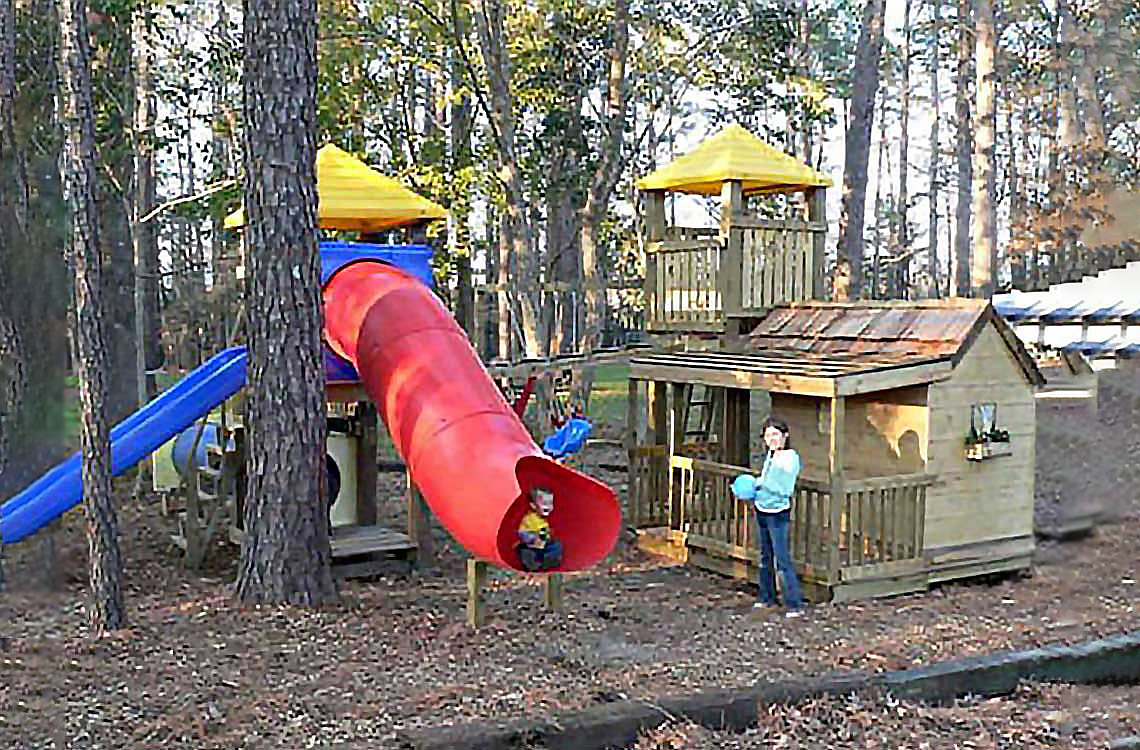 A young child emerges from a red tube slide as a woman looks on.