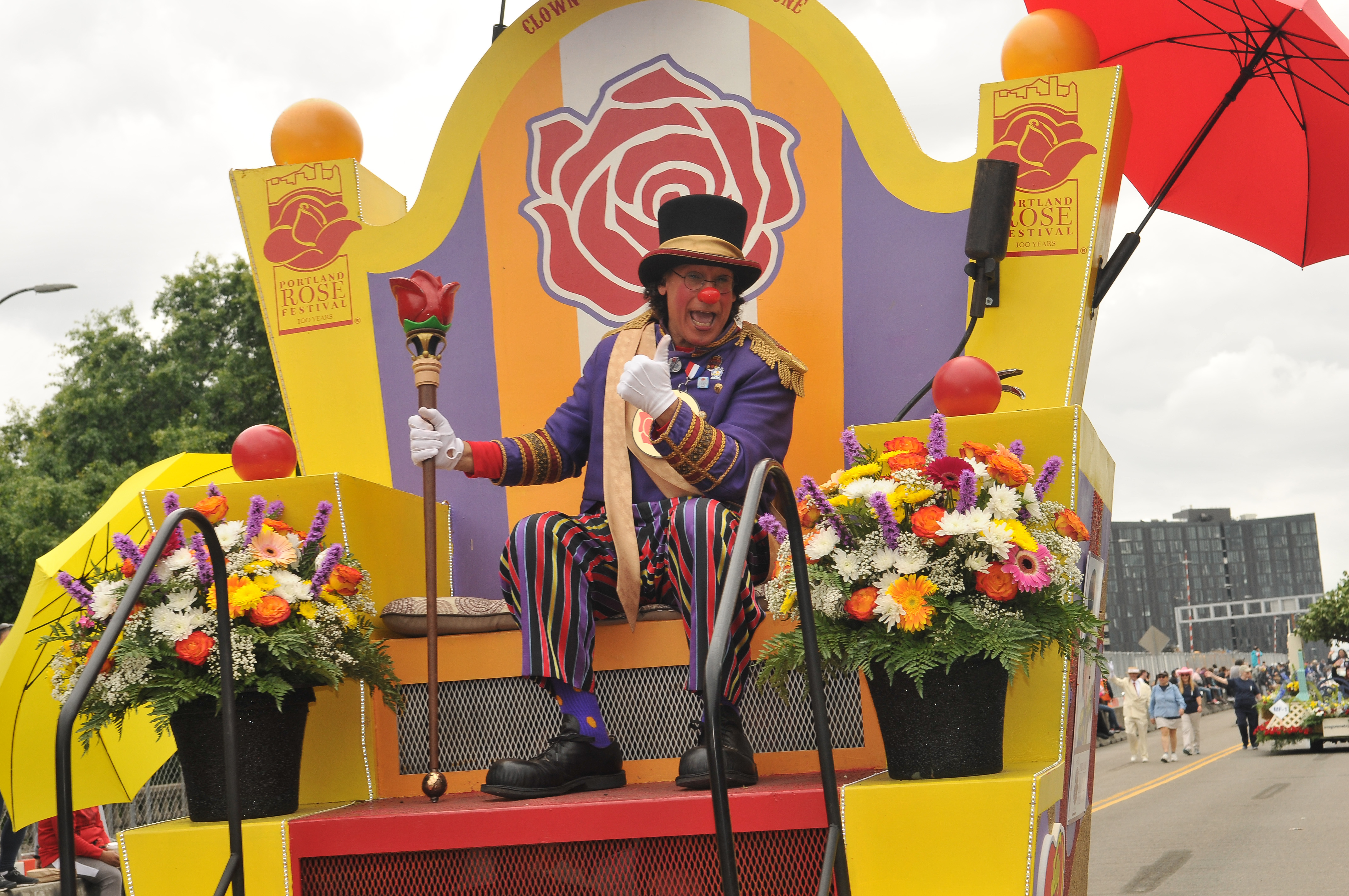 A man dressed as a clown sits on a throne mounted on a float rolling down a parade route.
