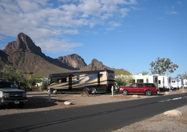 RVs parked on a dusty campground at the feet or rugged mountains.