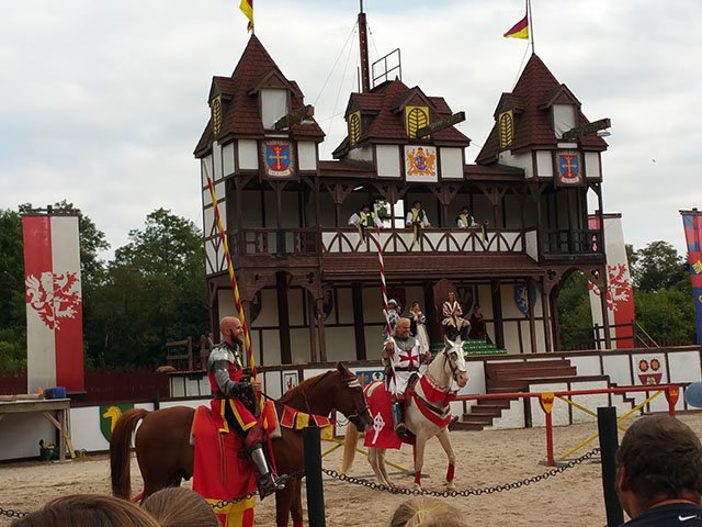 Men dressed in medieval garb on horseback with joisting poles in front of a Tudor castle.