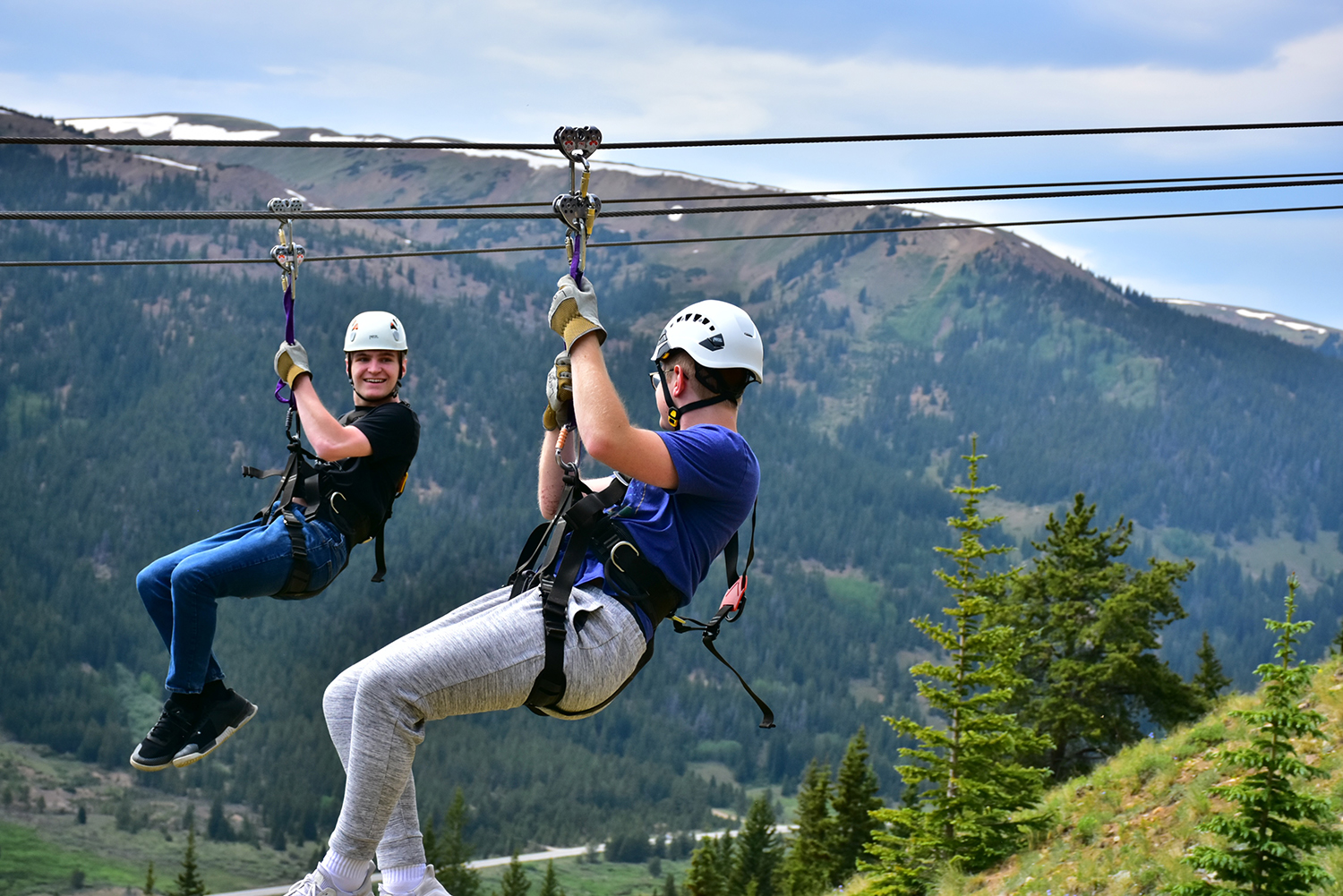 A pair of young men zooming down a zipline against a background of forested mountains.