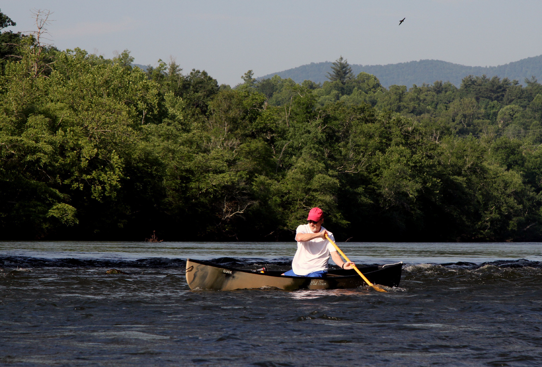 A lone canoer paddles on a river against a forested and mountainous background.