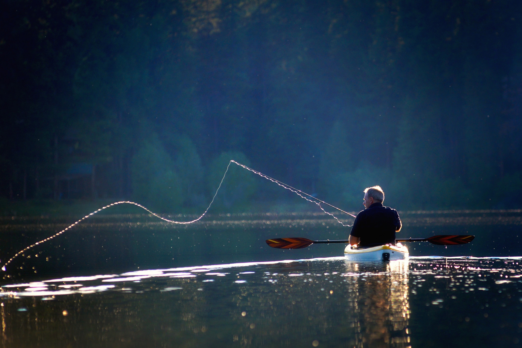 A fisherman casting on a lake from a kayak, backlit by the afternoon sun