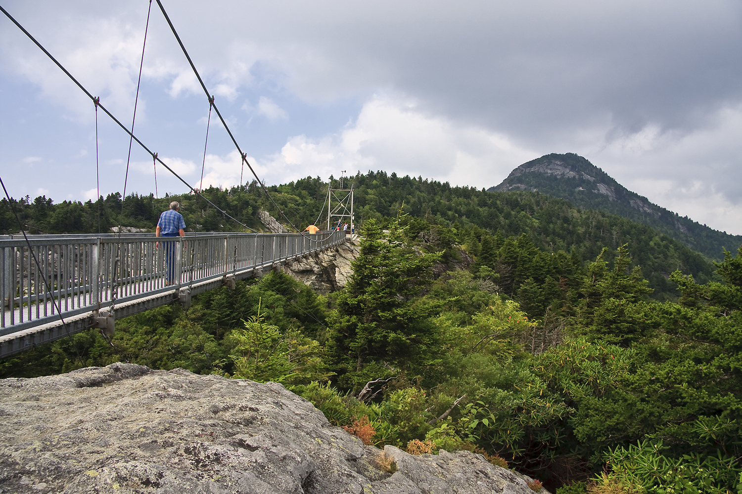 Hikers cross a suspension cable bridge toward a mountain in the distance.