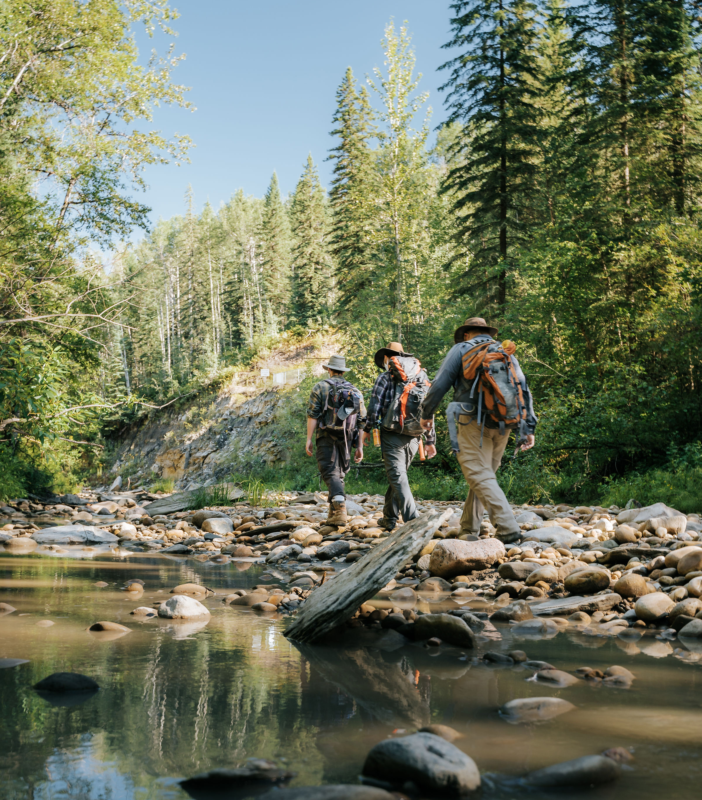 Three hikers walking on the rocky banks of a placid creek.