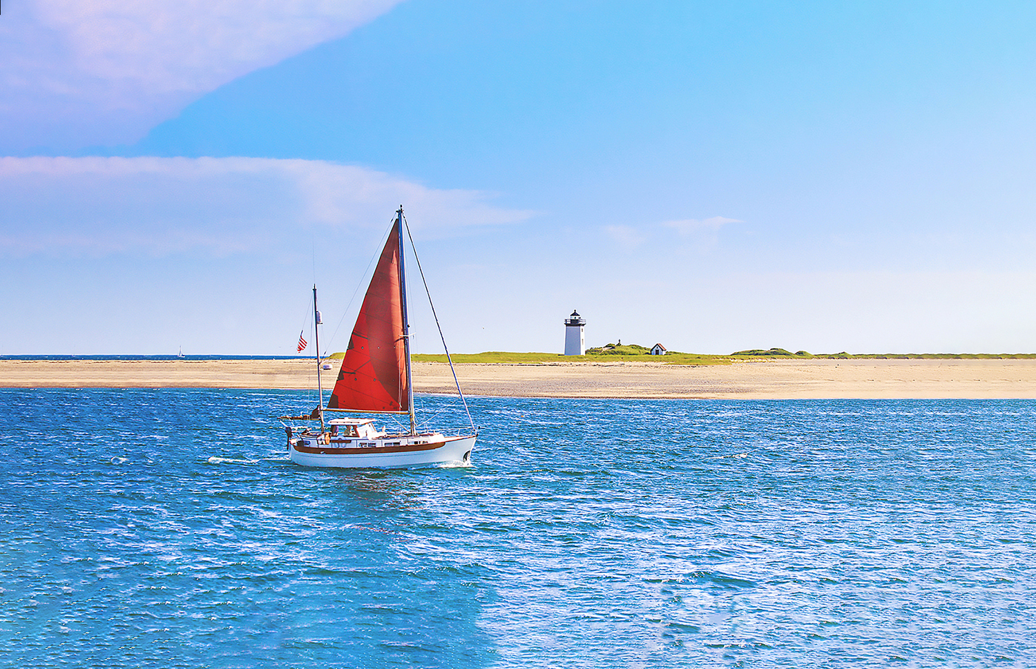 A red sailboat follows a course parallel to a sandy beach.