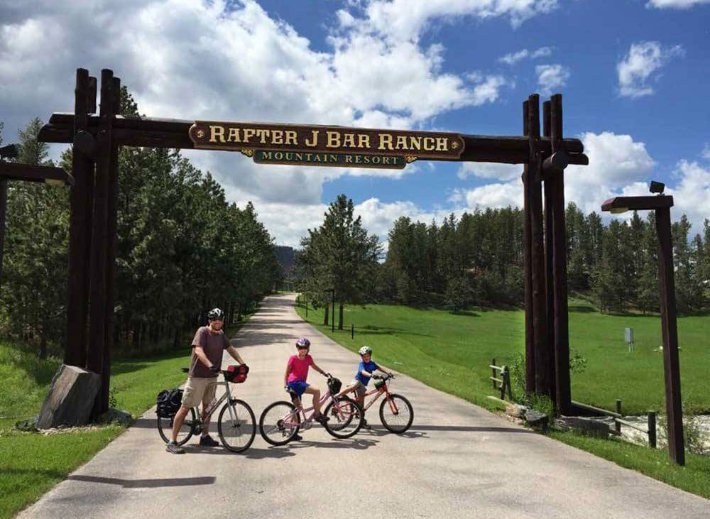 Three cyclists posing at the gate of an RV campground.